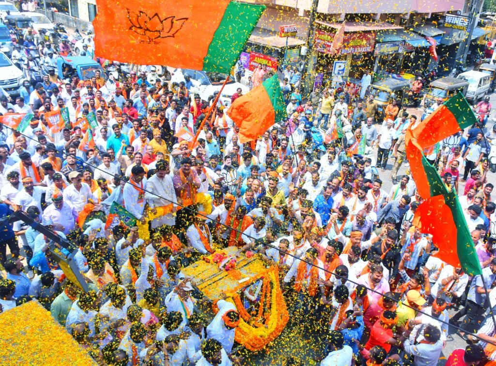 Crowds gather in Mangaluru for a colorful street parade with vibrant flags and decorations.